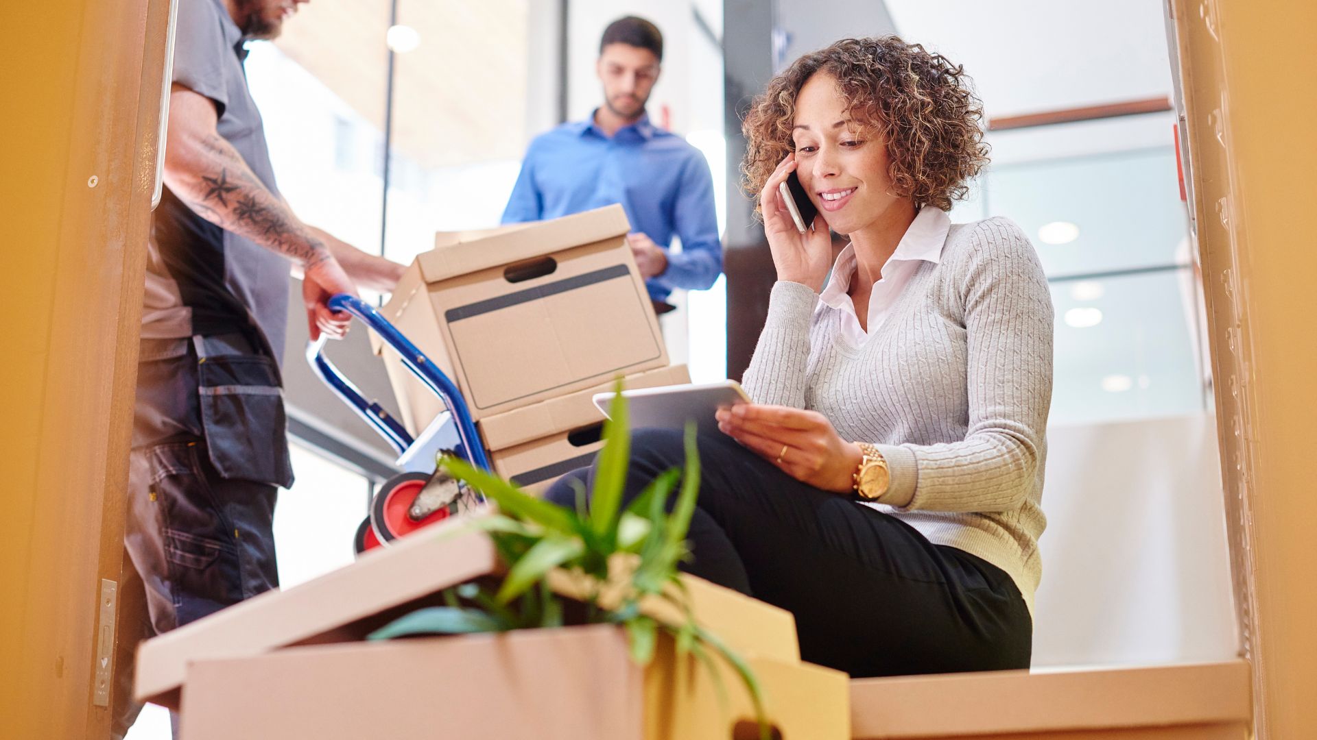 Woman sitting on stairs surrounded by moving boxes, talking on the phone and using a tablet during an office relocation.