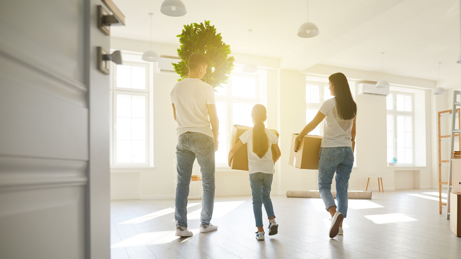 A family of three carrying boxes and a potted plant into a bright, empty room with large windows and light fixtures.