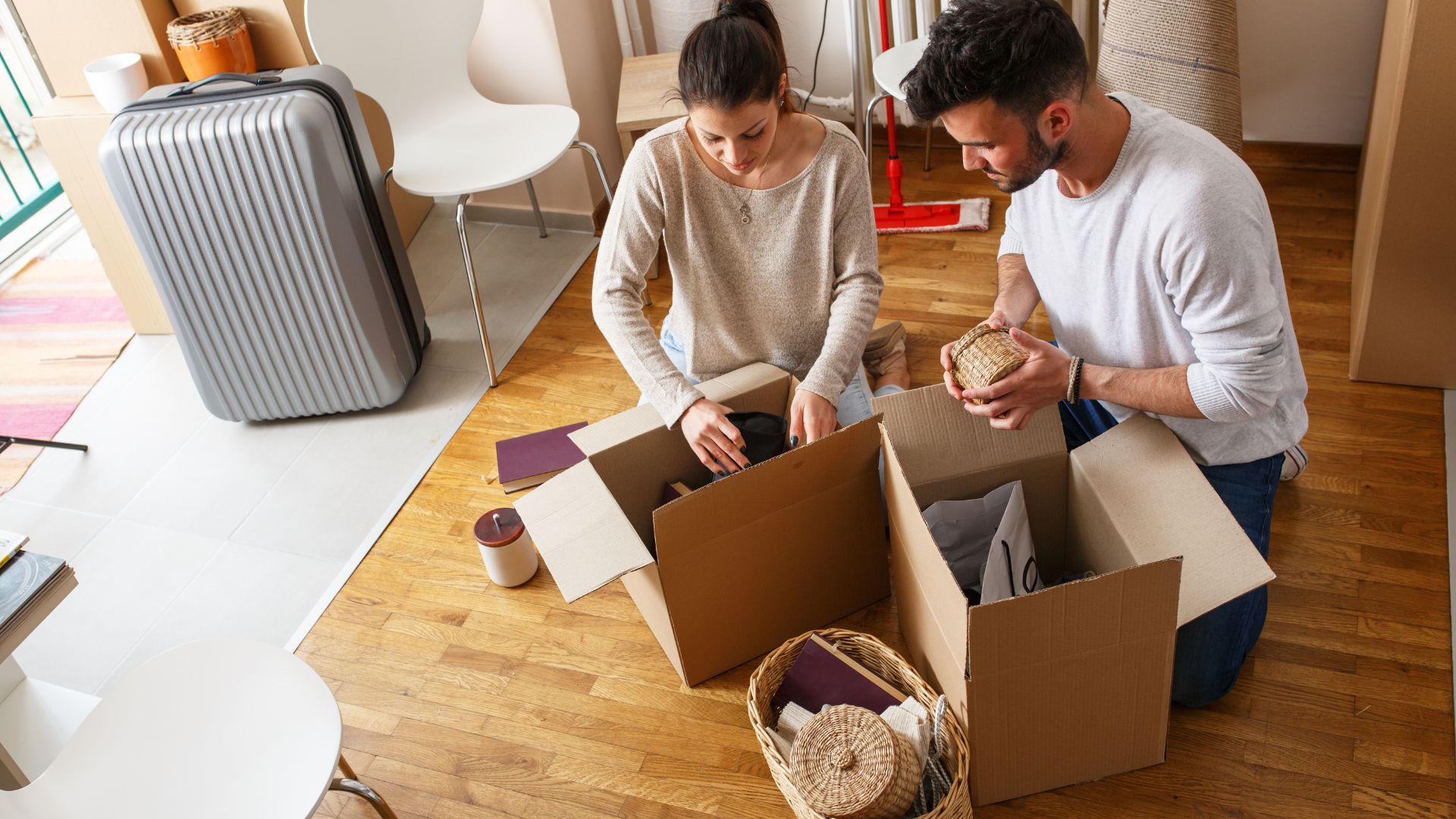 Young couple sitting on the floor of a partially packed room, organizing items into cardboard boxes surrounded by furniture and a suitcase.