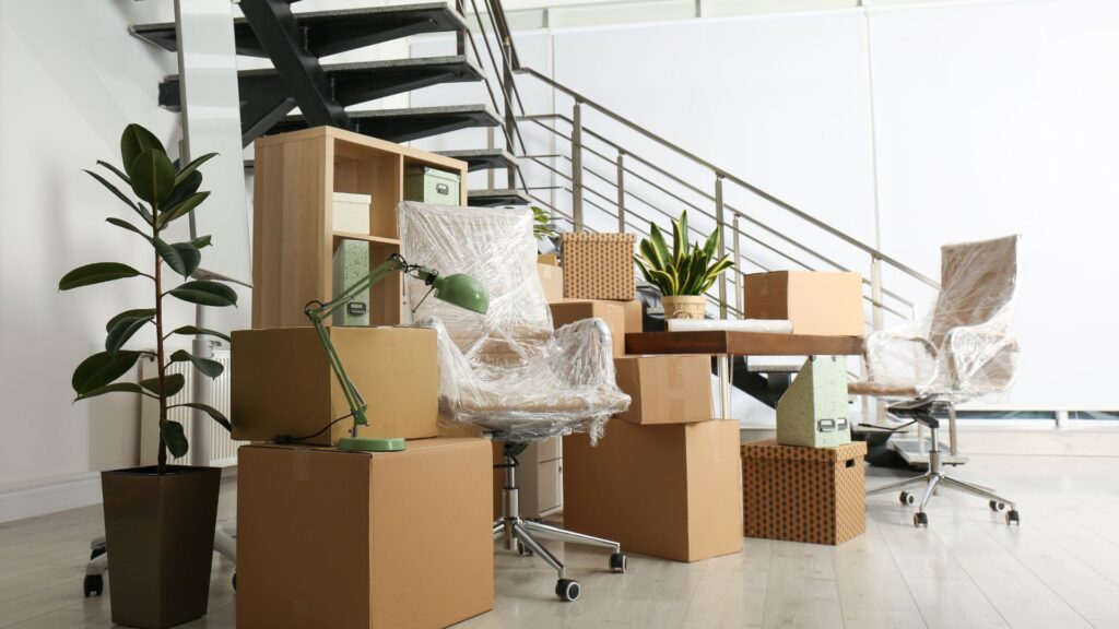 Office furniture wrapped in plastic and packed boxes arranged near a staircase in preparation for an office move.