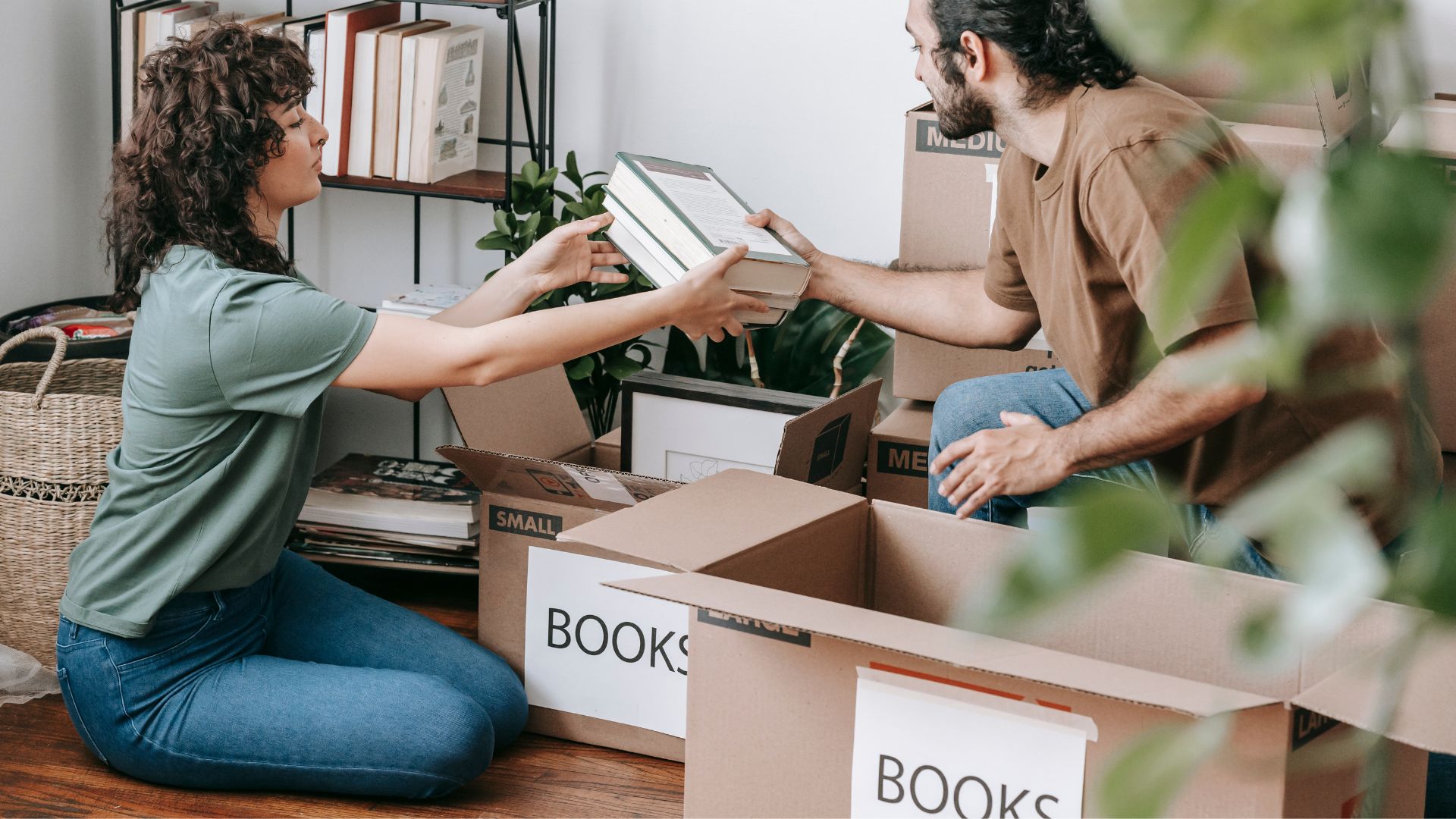 Man and woman packing labeled boxes with books in a cozy room, with a bookshelf and woven baskets in the background.