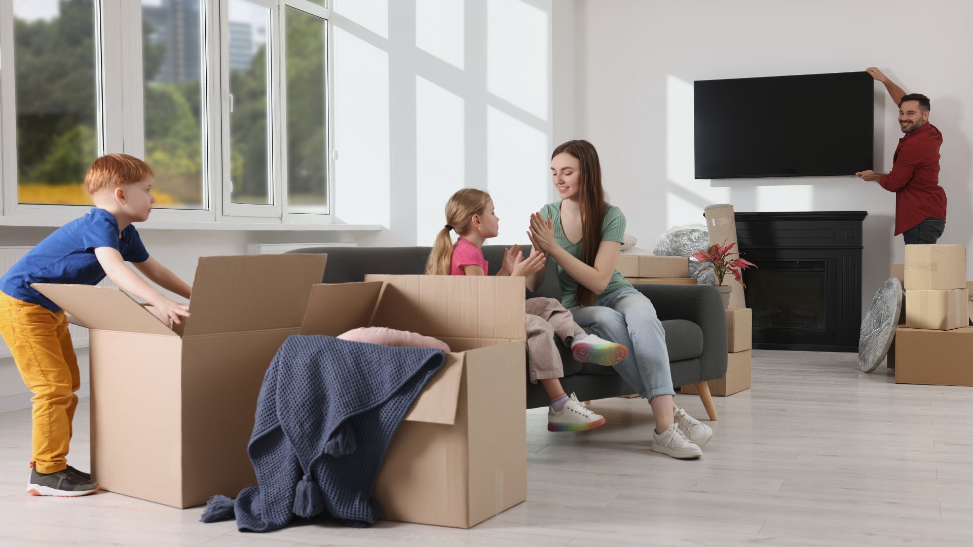 Family with young children unpacking moving boxes in a living room; the mother and daughter sit on the couch while the father installs a TV.